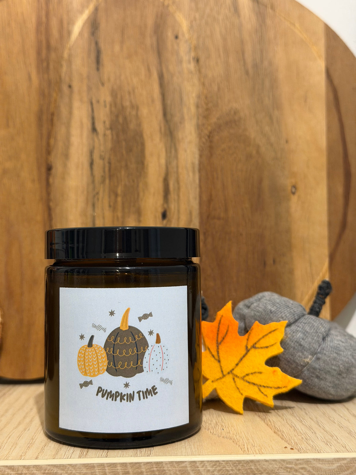 Pumpkin Spice szójagyertya barna üvegtégelyben, „Pumpkin time” címkével, kerek fa deszka előtt, mellette sárga filc juharlevél és szürke textiltökök