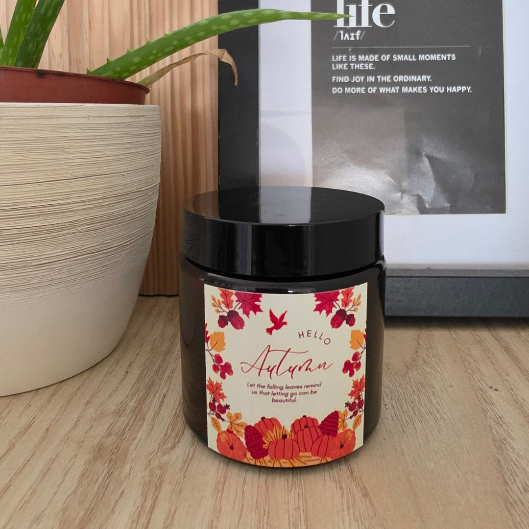 Fekete tetővel lezárt Pumpkin Spice szójagyertya barna üvegtégelyben, „Hello Autumn” feliratú, őszi virágkoszorús címkével, fa polcon, mellette aloe vera cserépben és keretezett kép