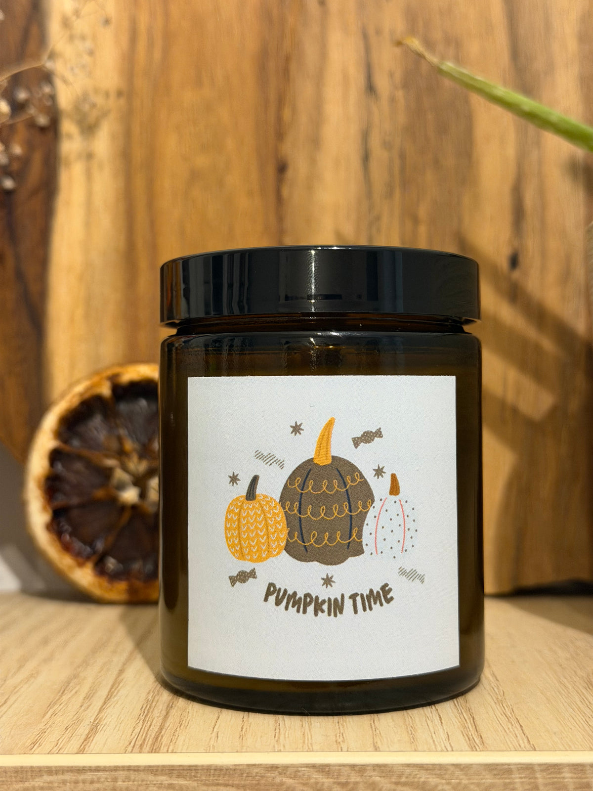 Fekete tetővel lezárt Pumpkin Spice szójagyertya barna üvegtégelyben, „Pumpkin time” feliratú, rajzolt tökös címkével, fa polcon, mellette szárított narancskarika