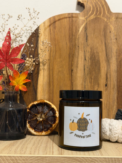 Pumpkin Spice szójagyertya barna üvegtégelyben, „Pumpkin time” címkével, tök alakú fa vágódeszka előtt, mellette szárított virágos váza, őszi levelek és fehér horgolt tök
