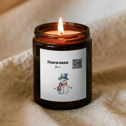 Égő Kiskedves „Dallamos” karácsonyi gyertya barna üvegtégelyben puha takarón, a címkén QR-kód, a Snowman felirat és akvarell hóember